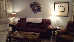 Un salon avec un canapé en cuir marron, un fauteuil blanc, une table basse et une table d’appoint avec une lampe.