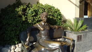 Une statue de bronze représentant une figure assise portant un nœud papillon, placée sur une zone pavée, avec un mur de pierre et un feuillage verdoyant en arrière-plan.