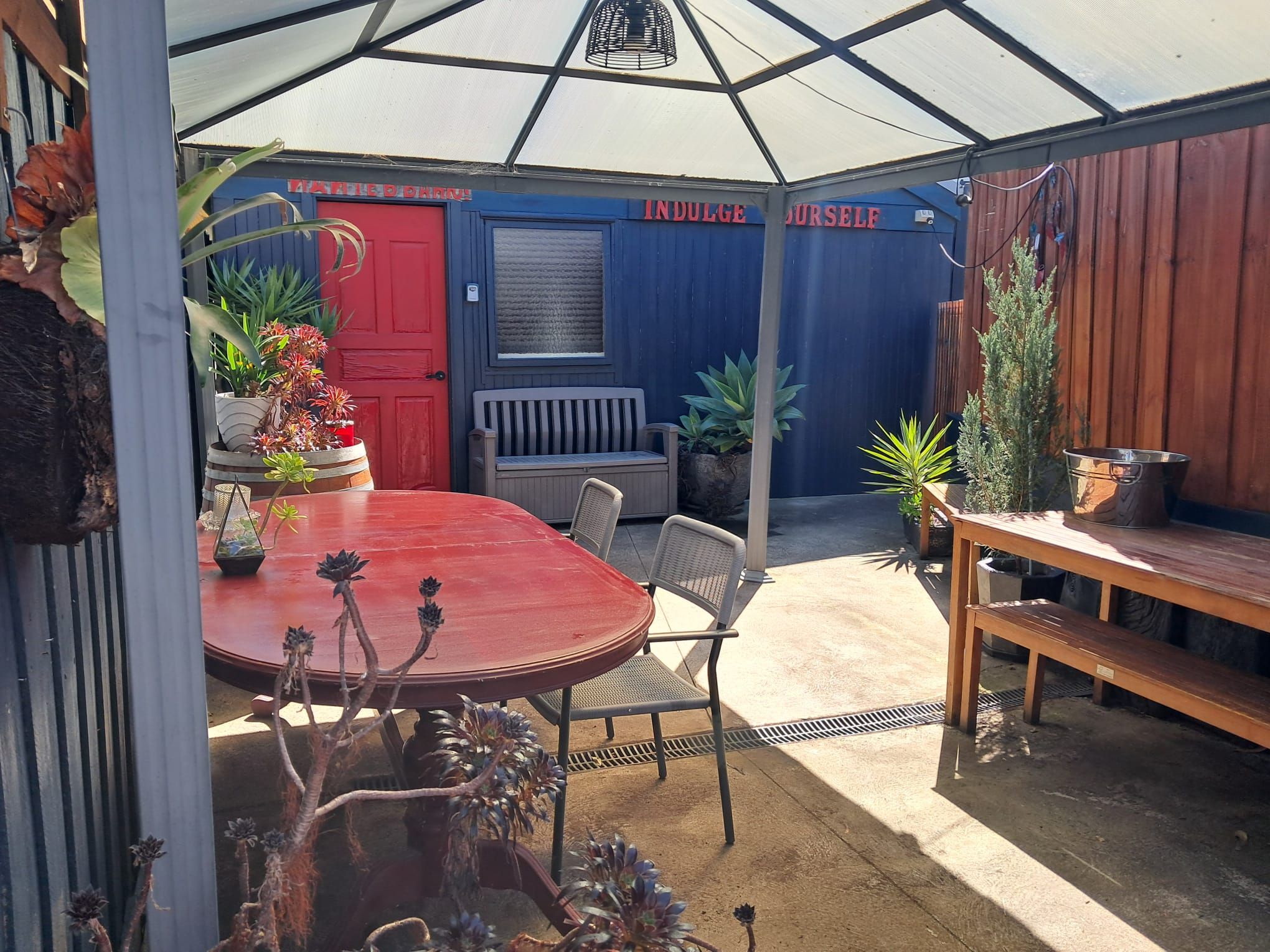 Un patio techado al aire libre con una mesa y sillas rojas, un banco de madera y diversas plantas en macetas.
