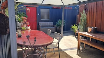 Un patio techado al aire libre con una mesa y sillas rojas, un banco de madera y diversas plantas en macetas.