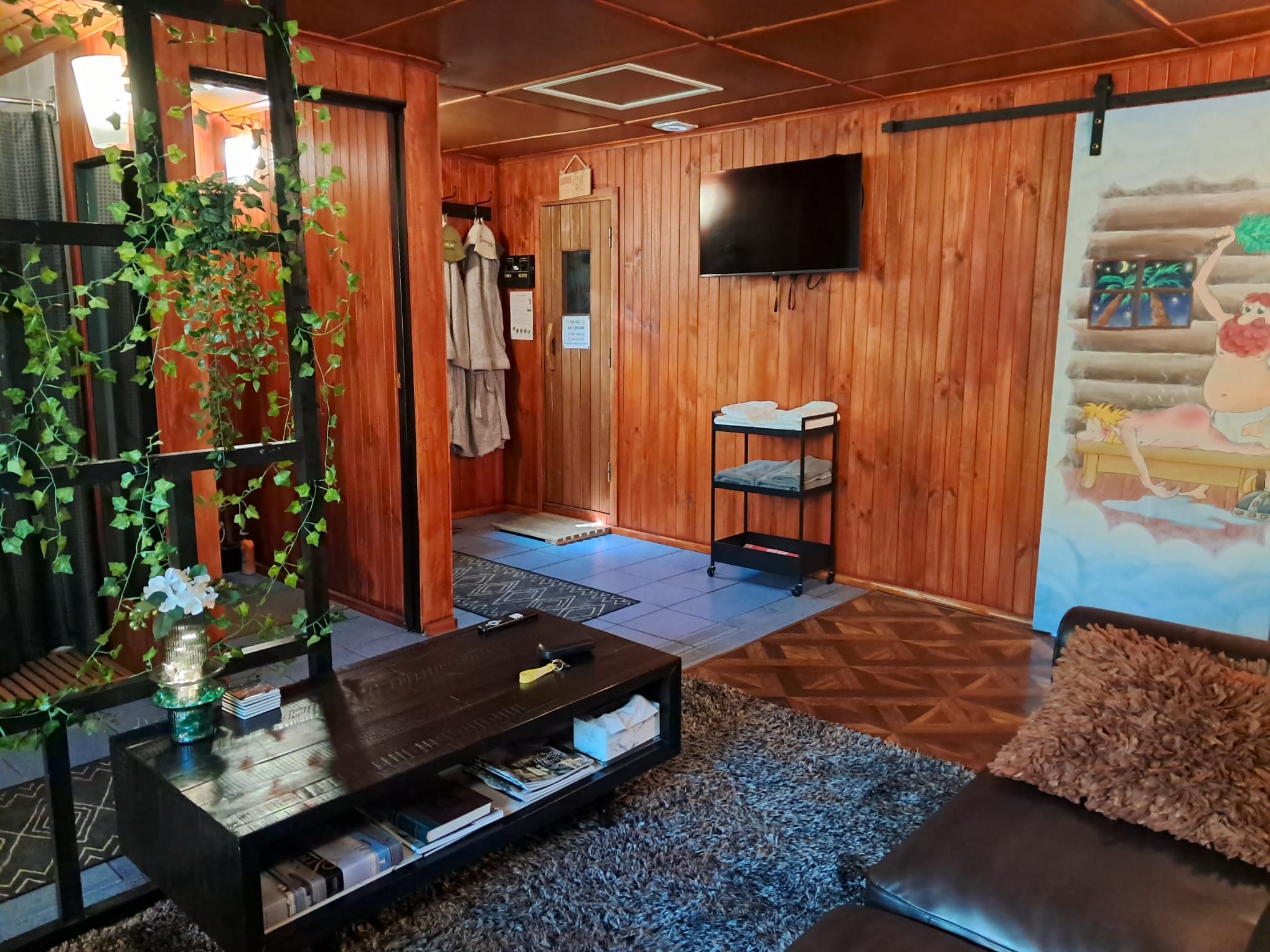 A room with wooden walls, a television mounted on the wall, a sofa, a coffee table with books, and a painting on the wall.