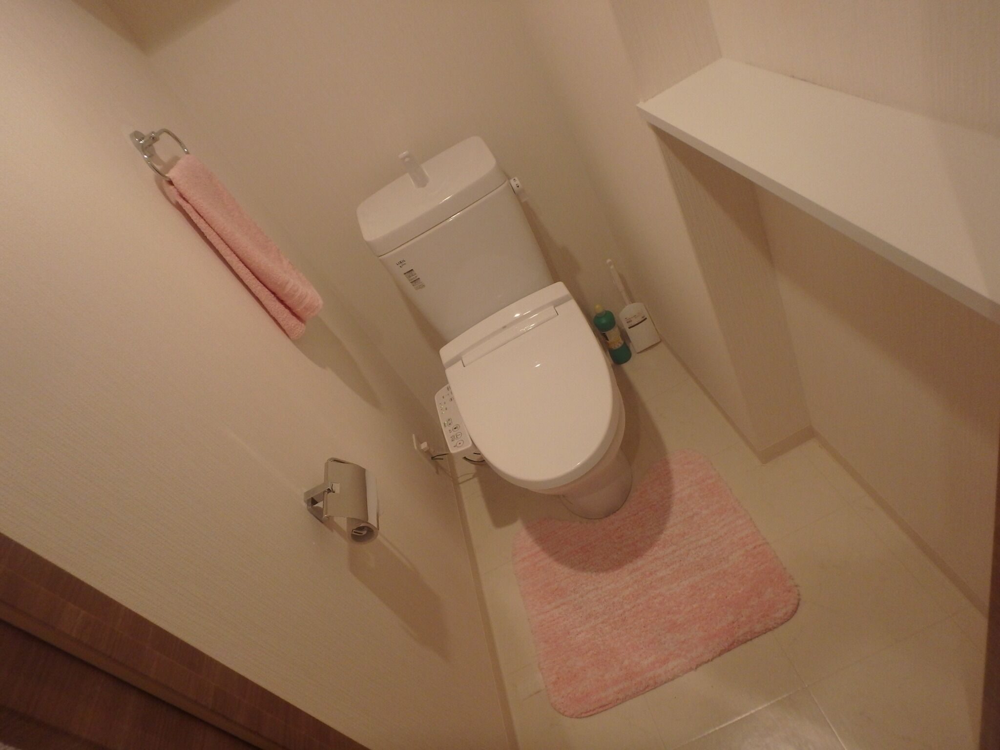 A bathroom with a toilet, a pink towel, a toilet paper holder, and a shelf.