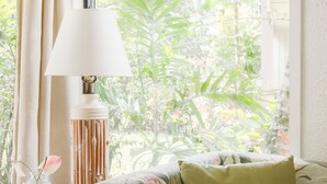 A room with a floral sofa, a table with a lamp, and a view of greenery outside. - Quaint stand-alone cottage - 2 bedroom 2 bath (Princeville)