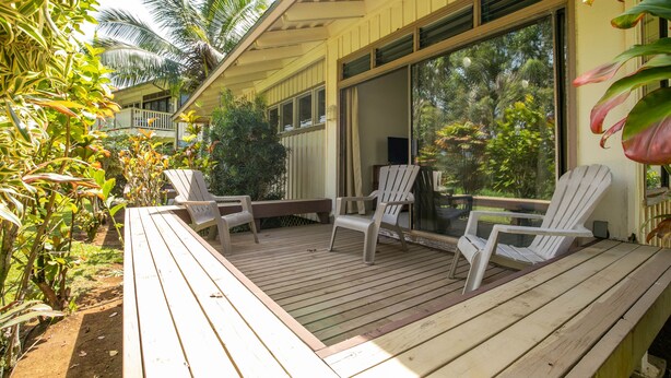 A wooden deck with white chairs and a bench, surrounded by tropical plants and a large window. - Quaint stand-alone cottage - 2 bedroom 2 bath (Princeville)