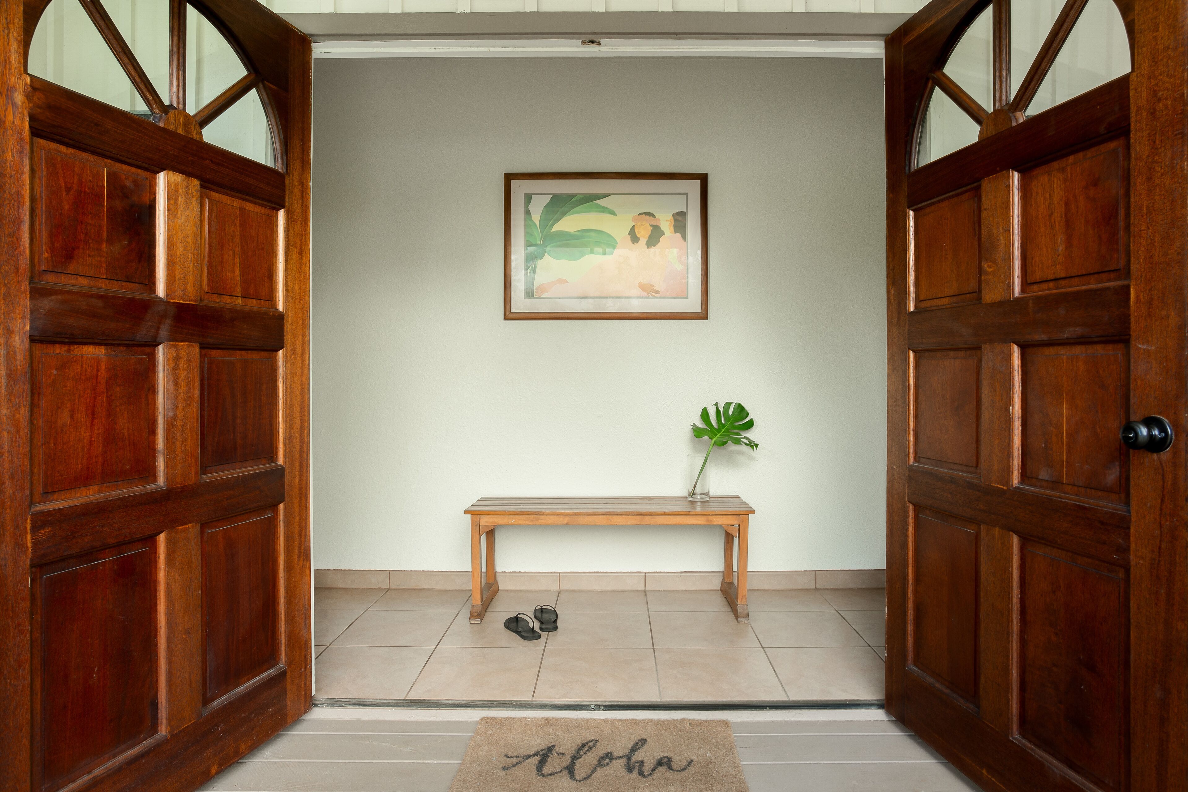 Puerta de madera con felpudo que dice "Aloha" que da a un espacio con un banco, un cuadro en la pared y una planta en maceta.