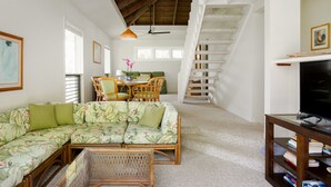 A spacious living area with a sectional sofa, a dining table with chairs, a staircase, and a television on a stand. - Quaint stand-alone cottage - 2 bedroom 2 bath (Princeville)