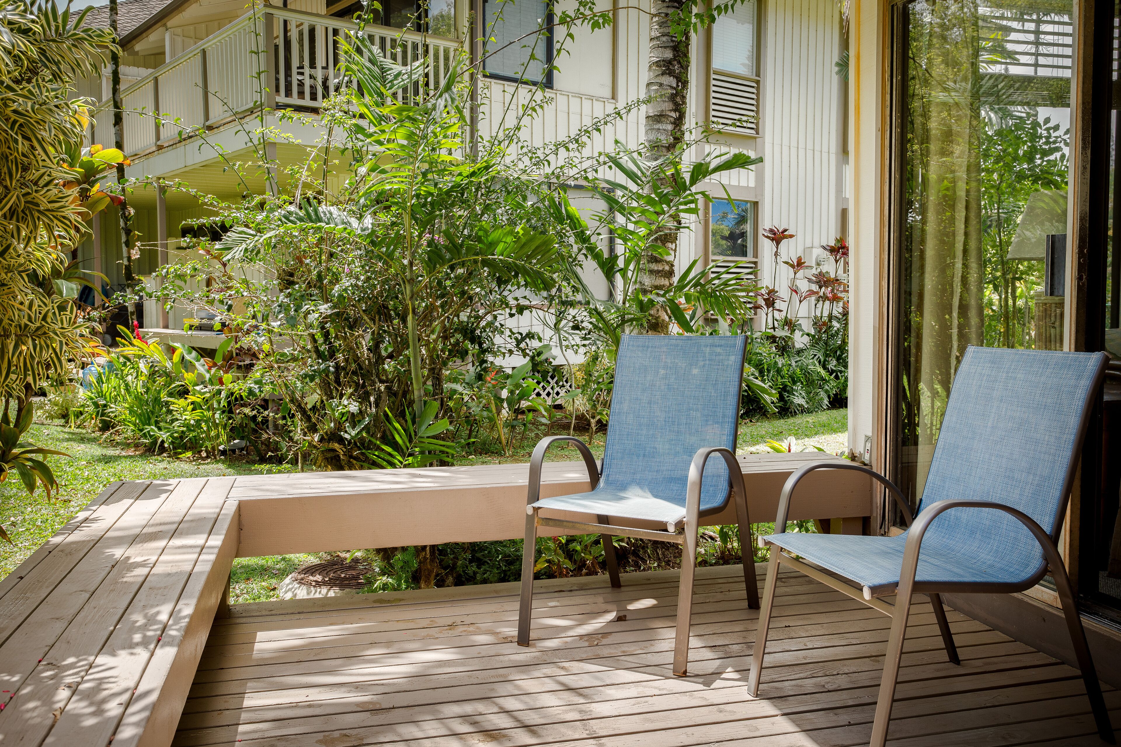 A wooden deck with two blue chairs and a table, surrounded by lush greenery and a building with a balcony.