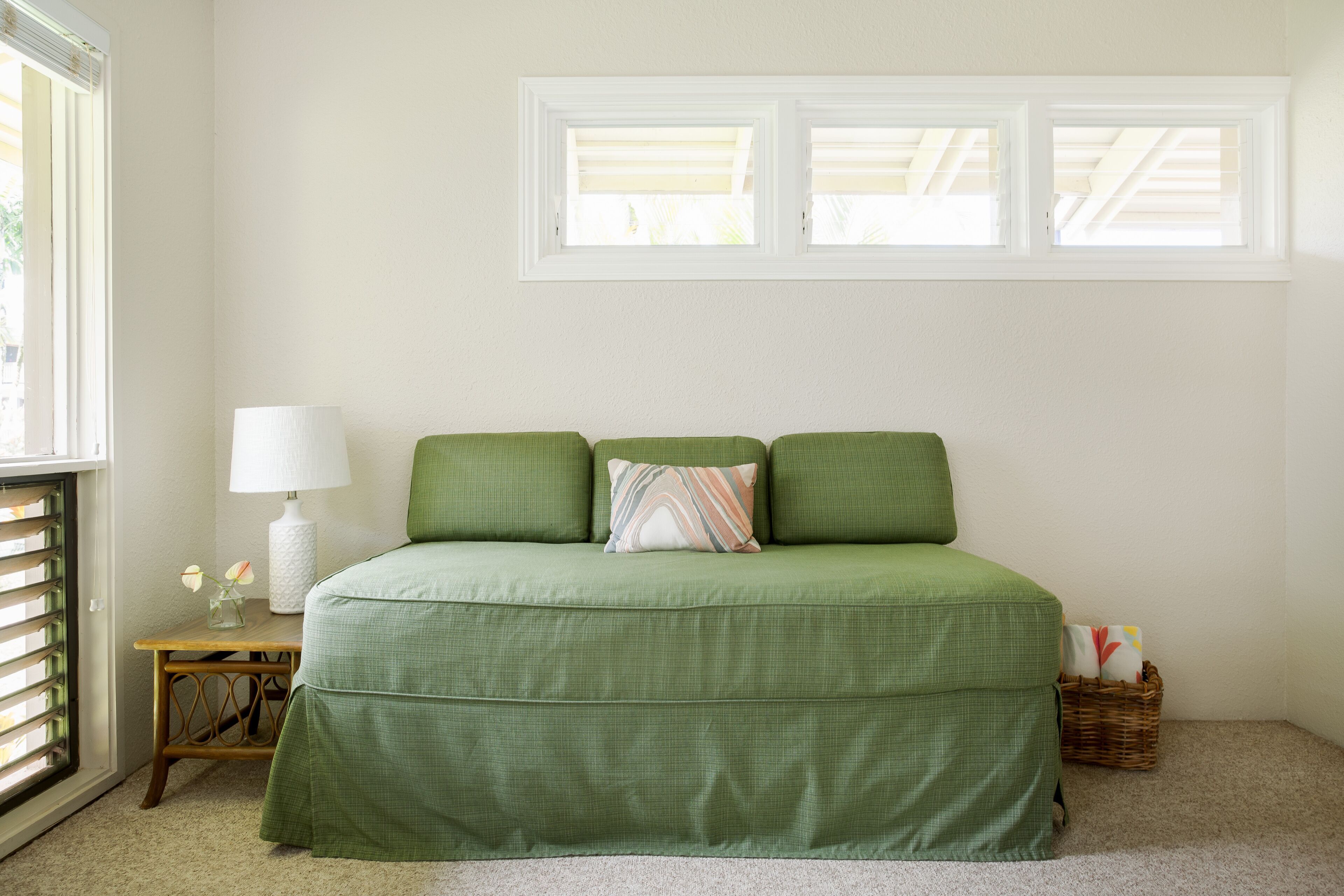 Una cama verde con una lámpara blanca y una mesita de noche, una ventana con persianas, y una cesta de mimbre en el suelo.
