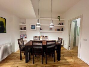 A dining area with a wooden table and chairs, pendant lights, white shelves with books and decorations, and a radiator on the floor.