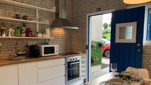 Une cuisine compacte avec des meubles blancs, un four à micro-ondes, un four traditionnel et un évier. Une porte bleue donne sur l’extérieur, et un petit coin repas est aménagé avec une table pour deux personnes.