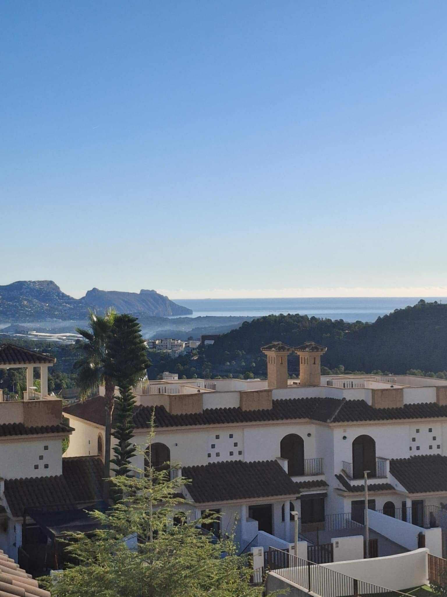A coastal town with terracotta-roofed buildings, a prominent tower, and a view of the sea and hills in the distance.