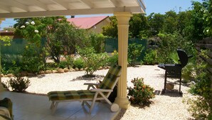 Een overdekt terras met een loungeruimte, een barbecue en een tuin met diverse planten en een hek.