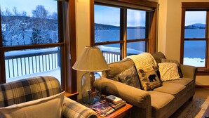 A cozy living room with a couch, a chair, a lamp, and a table with books. Large windows offer a view of a snowy landscape. - Rangeley Lake Lodge - On Rangeley Lake with large lawn and close to town! (Rangeley)