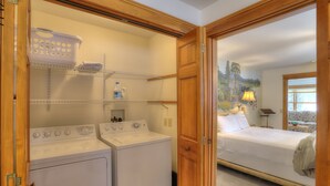 A laundry area with a washer and dryer, shelves with laundry baskets, and a bed in the adjacent room. - Rangeley Lake Lodge - On Rangeley Lake with large lawn and close to town! (Rangeley)