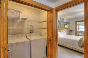Un área de lavandería con lavadora y secadora, repisas con cestos de ropa, y una cama en la habitación contigua.