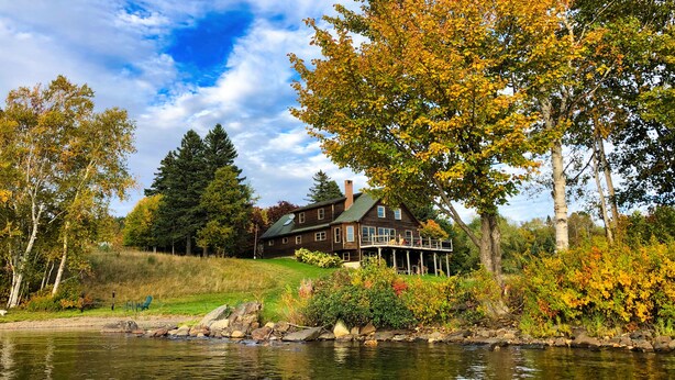 A large wooden house with a porch, situated by a lake, surrounded by trees and greenery. - Rangeley Lake Lodge - On Rangeley Lake with large lawn and close to town! (Rangeley)