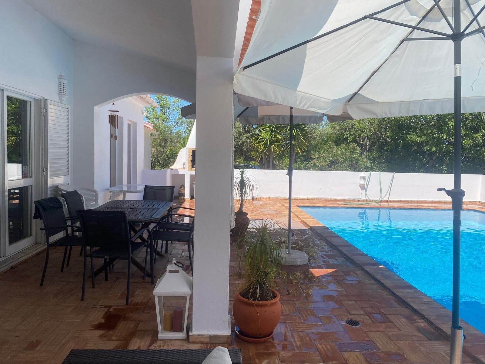 An outdoor patio with a pool, a table and chairs, a potted plant, and a white umbrella.