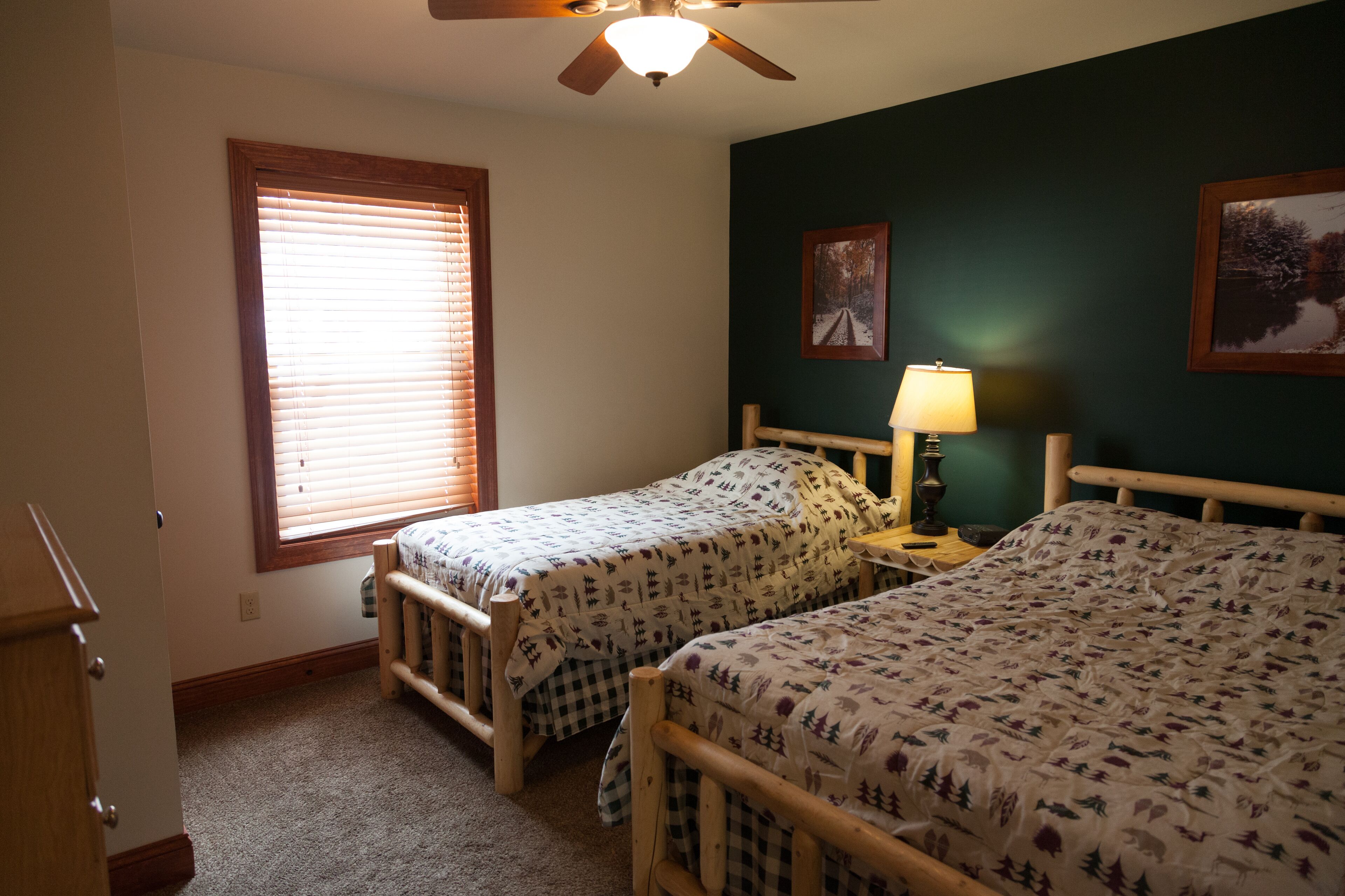 A room with two wooden beds, a ceiling fan, a window with blinds, and two wall-mounted lamps.