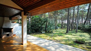 An open patio with a brick fireplace, a tiled floor, and a view of a forested area. - Seafront villa in Lignano Riviera (Lignano Sabbiadoro)