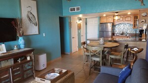 A living room with a sofa, a coffee table, and a dining area with chairs and a table. There is a kitchen area with cabinets and a refrigerator.