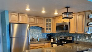 A modern kitchen with a stainless steel refrigerator, a microwave, an oven, and a herringbone-patterned backsplash. There are wooden cabinets, a granite countertop, and a stainless steel trash can.
