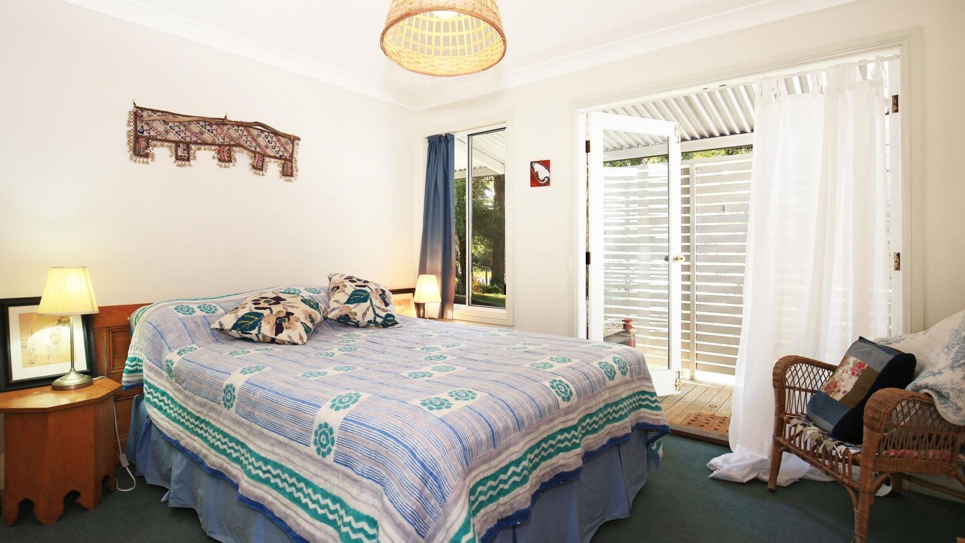 Un dormitorio con cama, mesita de noche, lámpara, silla de mimbre y vista al exterior a través de una puerta corredera.