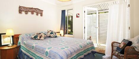 Un dormitorio con cama, mesita de noche, lámpara, silla de mimbre y vista al exterior a través de una puerta corredera.