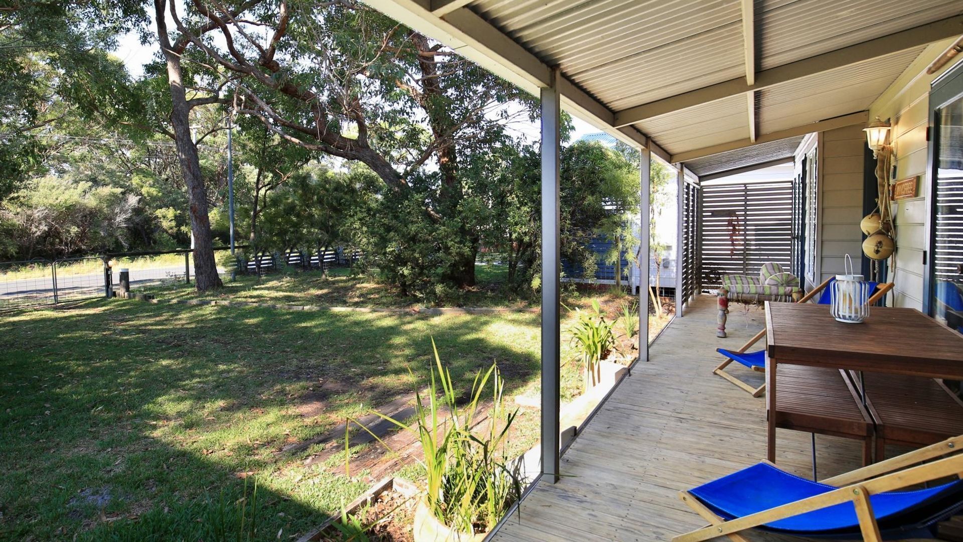 A covered outdoor patio with a wooden table and chairs, surrounded by greenery and trees.