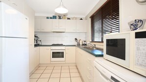 Une cuisine de taille réduite avec des meubles blancs, une cuisinière, un four, un four à micro-ondes et une fenêtre avec des stores.