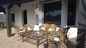 Une terrasse extérieure aménagée avec des meubles en osier et une table à plateau de verre, le tout adossé à un bâtiment blanc doté de grandes fenêtres.