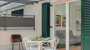 Un patio exterior con mesa y sillas, una parrilla y un cerramiento de vidrio corredera que da a una habitación interior con sofá y obras de arte.