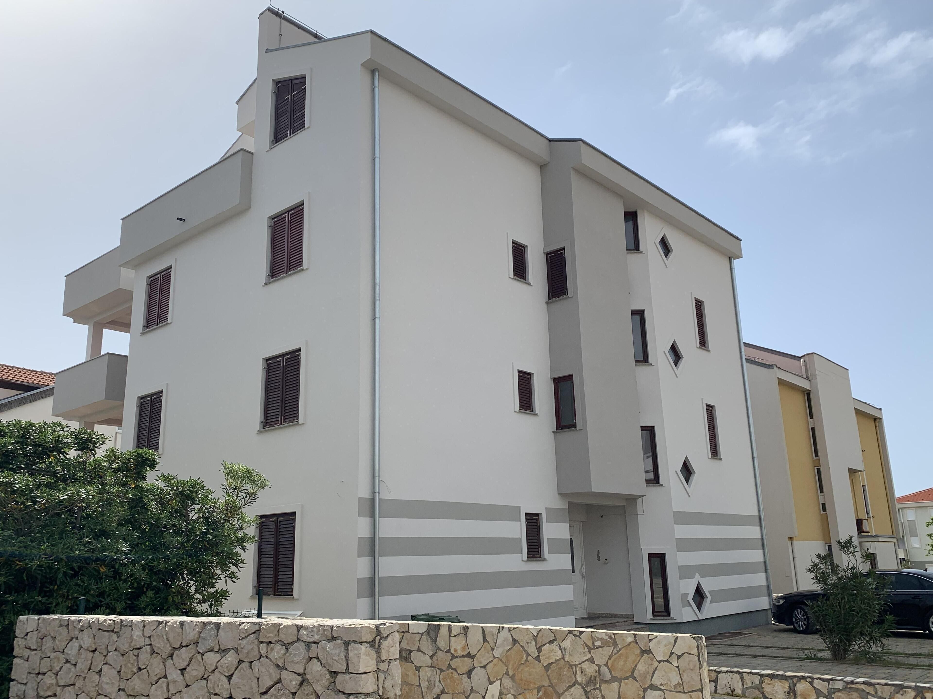 A modern multi-story building with balconies, a stone wall in the foreground, and a clear sky.
