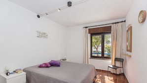 Un dormitorio con cama, mesita de noche, una silla, una ventana con cortinas y un reloj de pared.