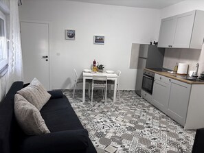 Ein kompakter Wohnbereich mit schwarzem Sofa, Esstisch mit Stühlen, einer Küchenzeile und einem Boden mit Muster.