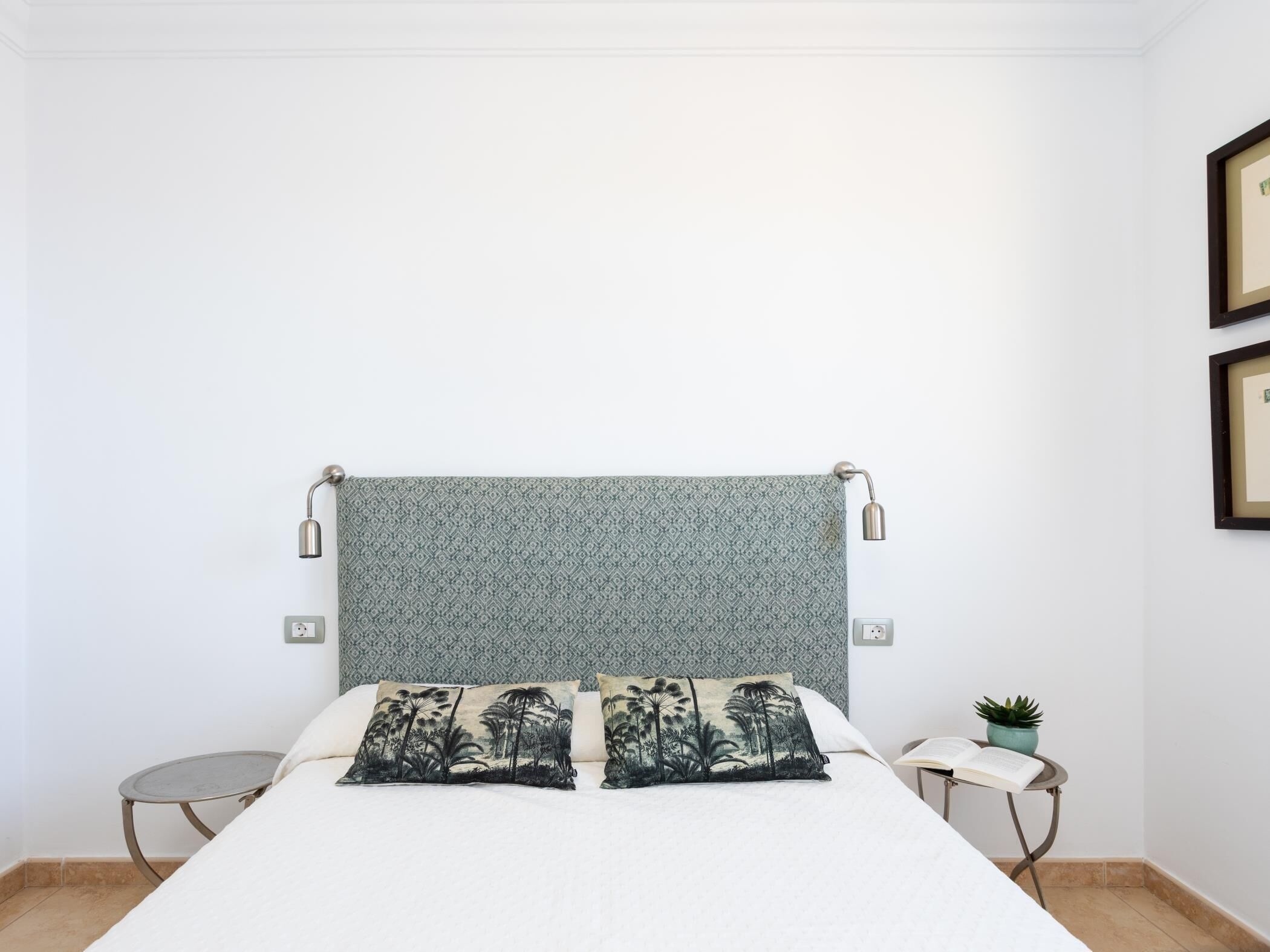 Une chambre à coucher avec un lit, deux oreillers, une petite table sur laquelle est posée une plante, et un luminaire fixé au mur.