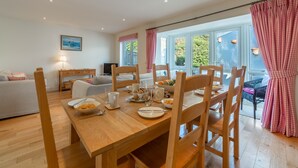 A dining area with a wooden table set for breakfast, surrounded by wooden chairs. A sofa and a TV are visible in the background. - No 4 Pentire Rocks is a contemporary maisonette situated in New Polzeath. (Wadebridge)