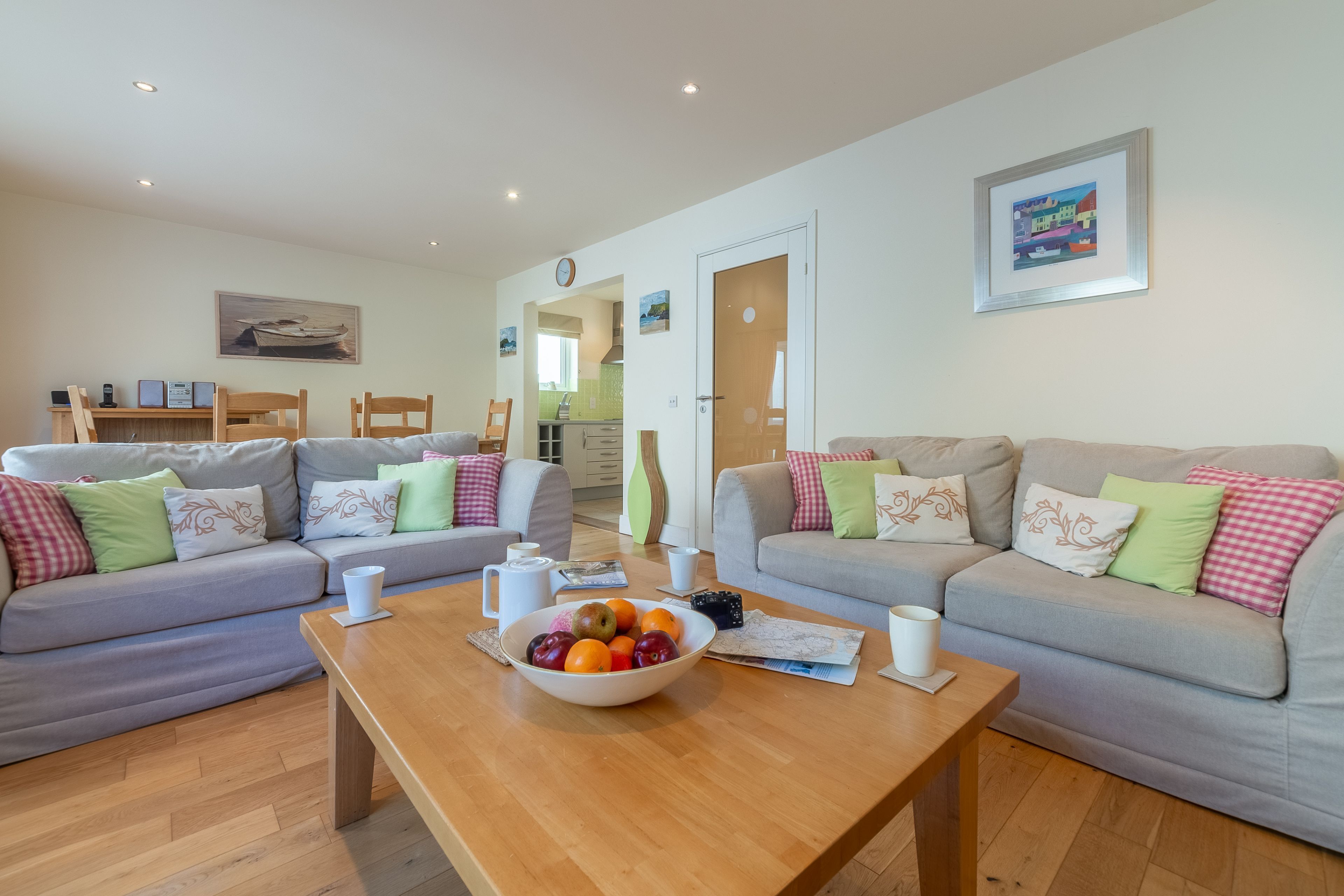 A modern living room with two grey sofas, a wooden coffee table with a fruit bowl, and a kitchen visible through an open door.
