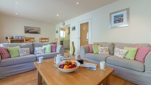A modern living room with two grey sofas, a wooden coffee table with a fruit bowl, and a kitchen visible through an open door. - No 4 Pentire Rocks is a contemporary maisonette situated in New Polzeath. (Wadebridge)