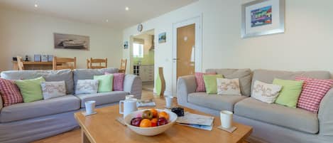 A modern living room with two grey sofas, a wooden coffee table with a fruit bowl, and a kitchen visible through an open door.