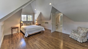 Un dormitorio con techo inclinado, una cama con cubrecama blanco, suelo de madera, una mesita de noche, una lámpara, una silla y una puerta que da al baño.