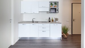 Une cuisine moderne avec des armoires blanches, un four à micro-ondes encastré et un évier. On aperçoit une plante sur le sol et une porte sur la droite.
