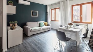 Un salon moderne avec un canapé gris, une table à manger avec des chaises, un petit coin cuisine et une grande fenêtre avec des rideaux.