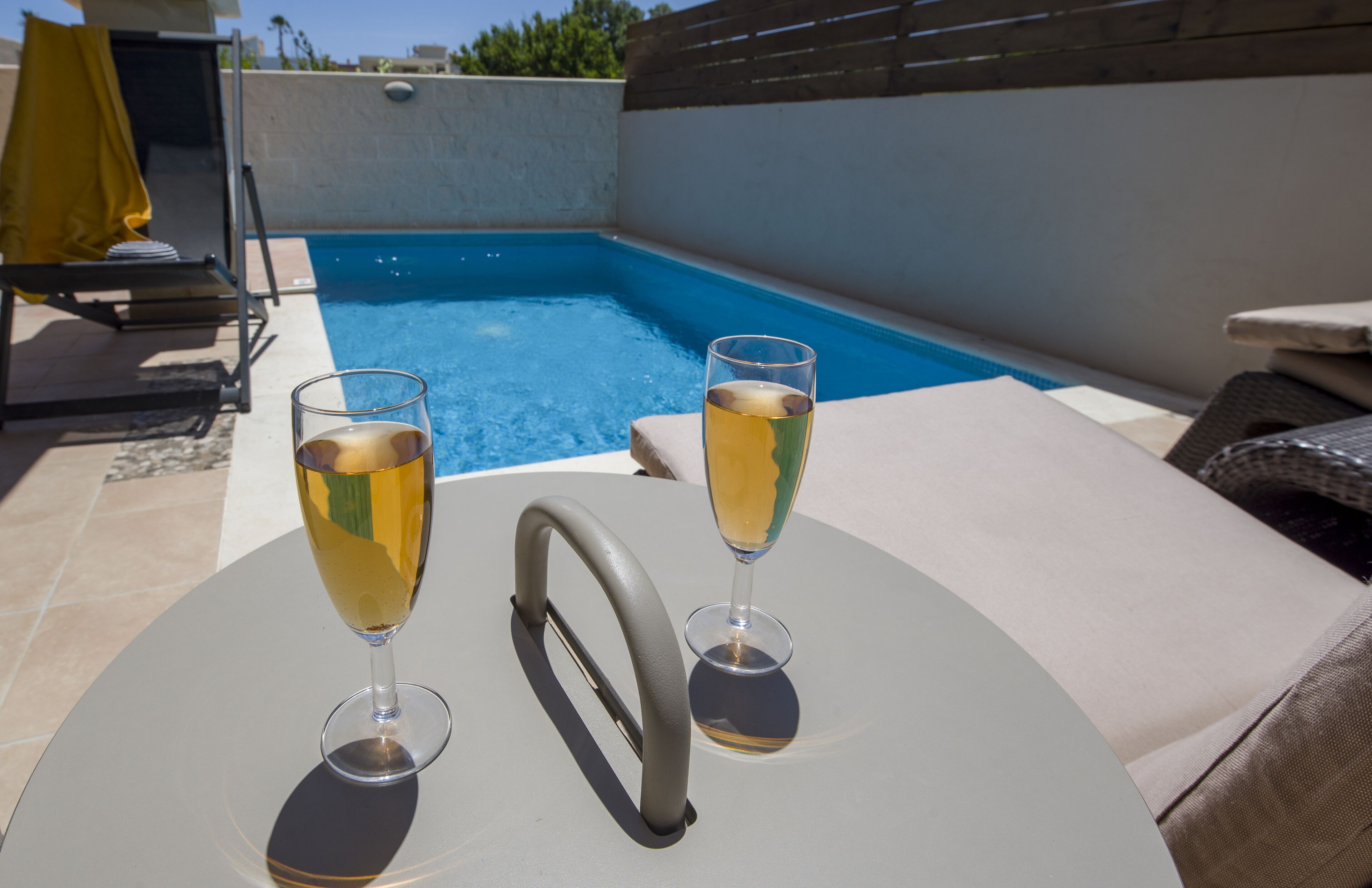 A poolside setting with two glasses of a clear beverage on a table, a sun lounger, and a pool.