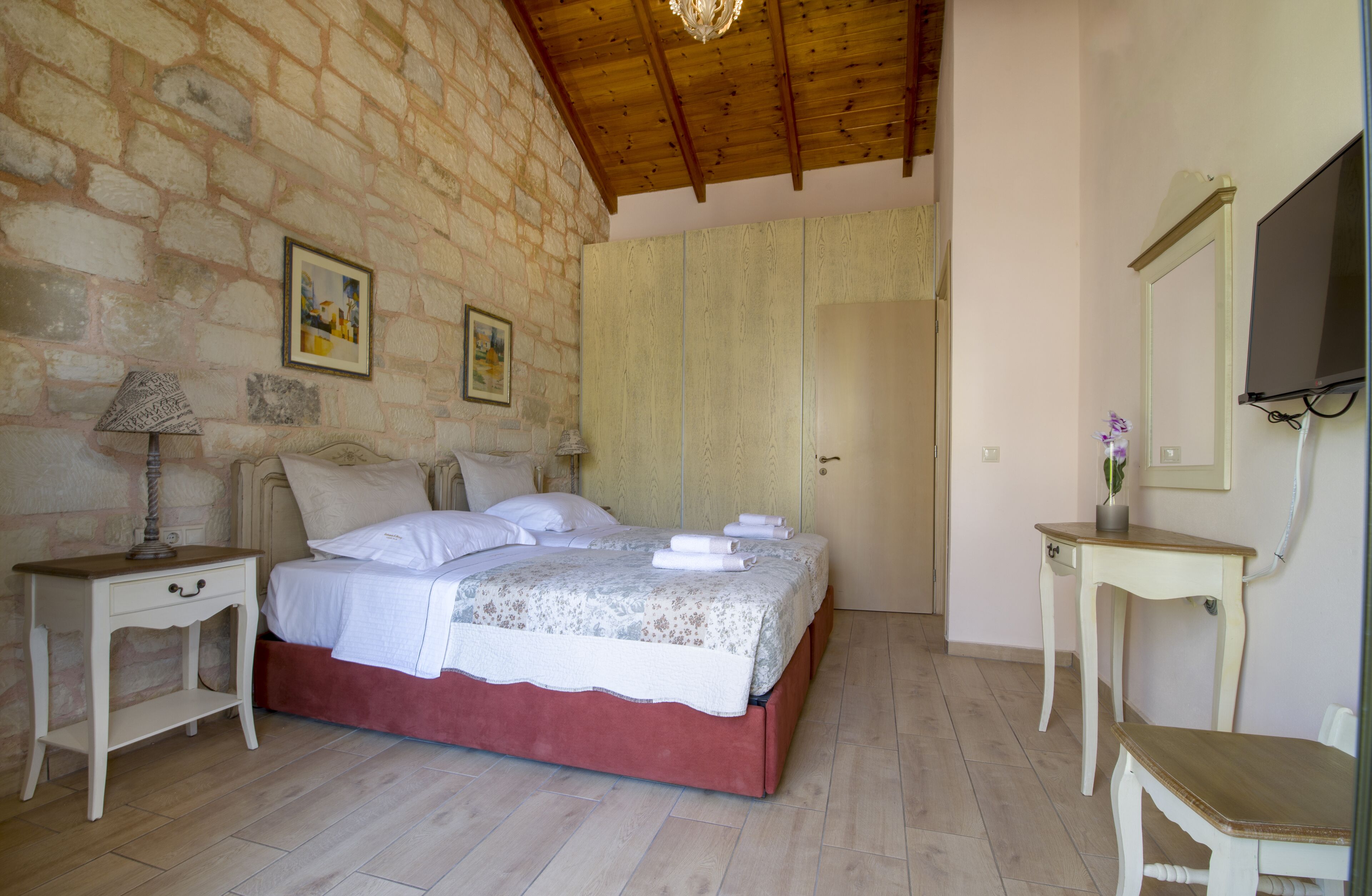 A bedroom with a stone wall, a wooden ceiling, a bed with white linens, a nightstand with a lamp, a bedside table, a mirror, and a door.
