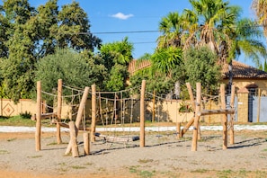 A fenced outdoor area with a climbing structure, surrounded by trees and a building with a tiled roof. - Cubo's Casa María los Javieles / free wifi / private pool (Alhaurín el Grande)
