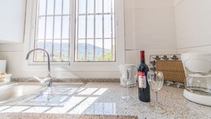 Una cocina con un lavamanos, dos ventanas, una botella de vino y una cafetera.