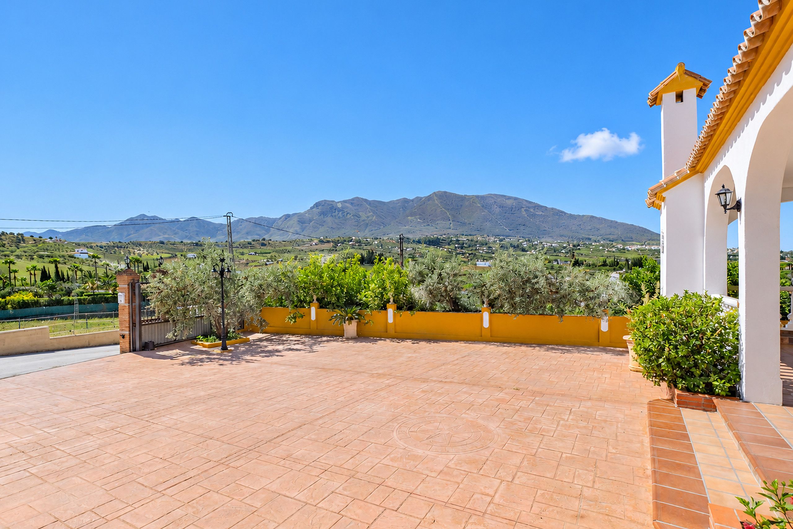 A terrace with a view of a mountain range, a yellow wall, and a white building with a tiled roof.
