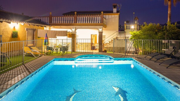 Una piscina con borde de baldosas, un porche techado y un edificio de dos pisos con balcón.