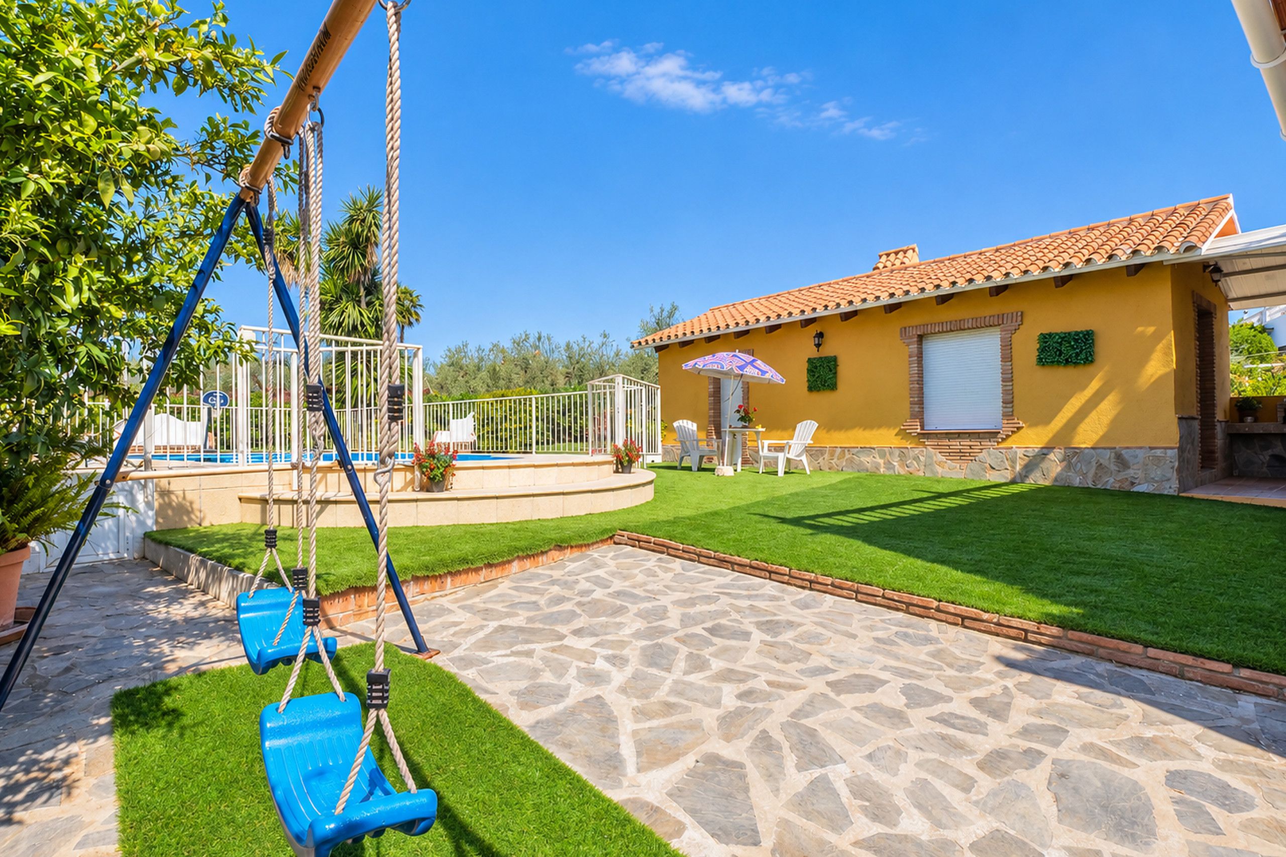 A patio with a swing set, a small pool, and a yellow building with a tiled roof.