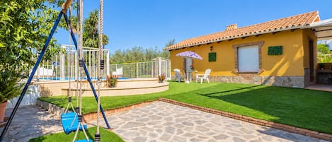 A patio with a swing set, a small pool, and a yellow building with a tiled roof.